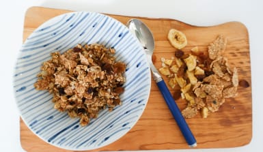 Eine blau-weiße Schale gefüllt mit Müsli steht auf einem Olivenholzbrett, daneben liegen ein Esslöffel, Bananenchips und Flakes.